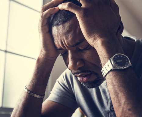 The man is wearing a gray shirt, accessorized with a wristwatch and bracelet, sitting in a dimly lit space near a window. His body language, with hands cradling his head and eyes closed, emphasizes emotional turmoil, perhaps due to personal, financial, or mental health challenges. The lighting adds to the mood of distress or reflection.