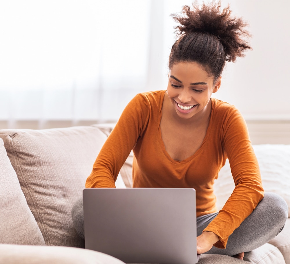 The woman is casually dressed in a burnt orange shirt and gray leggings, sitting cross-legged on a beige couch in a well-lit living room. Her expression is joyful and focused, implying she is engaged in a positive or productive online activity, such as learning, working, or connecting with others.