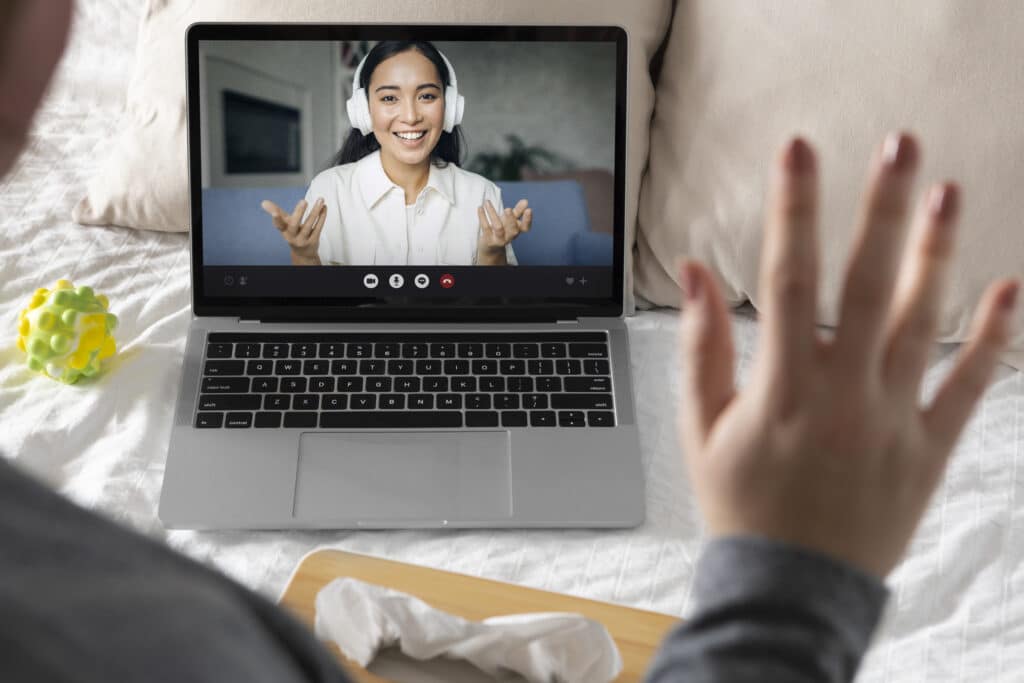 A laptop displays a video call with a smiling woman wearing white headphones, gesturing enthusiastically. The laptop is placed on a bed, surrounded by casual items like a toy and tissues, indicating a relaxed or informal virtual interaction. A hand is raised in a waving gesture in the foreground.