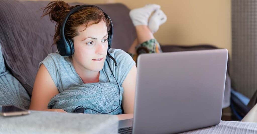 Teenage girl or young adult with light skin and brown hair, wearing a black over-ear headset, lying on a gray couch. She is propped up on a blue textured pillow, looking intently at a silver laptop. She wears a light gray t-shirt and patterned shorts, with white socks visible as her feet are raised behind her. The setting appears casual and comfortable, suggesting a relaxed home environment during a teletherapy session.