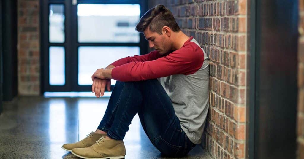 Teen boy sitting alone on the floor in a hallway, leaning against a brick wall with his head down, appearing distressed.