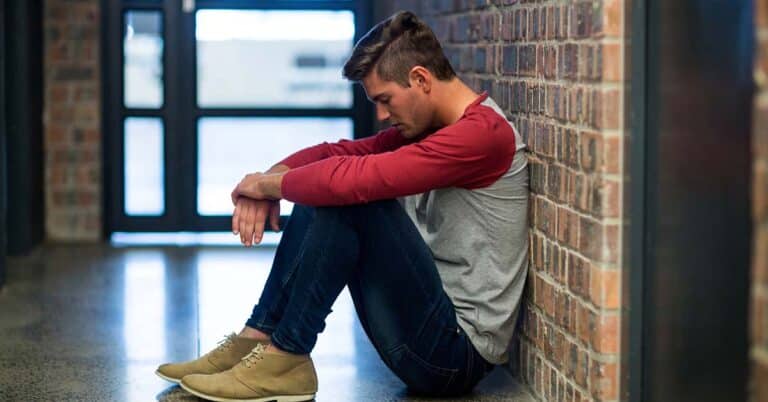 Teen boy sitting alone on the floor in a hallway, leaning against a brick wall with his head down, appearing distressed.
